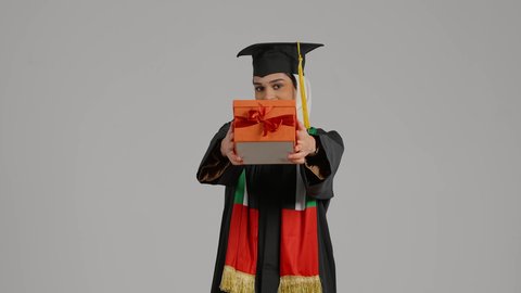 النظر الى الكاميرا بايماءات السرور،الاحتفالات و المناسبات الجامعية, جودة التعليم في المملكة، الشعور بالفخر والثقة بالنفس، إنهاء الدراسة الجامعية، مفهوم الاجتهاد والتفوق، تصوير مقرب لطالبة عربية خليجية اماراتيه محجبة ترتدي عباءه وقبعة التخرج تحمل بيدها هدية النجاح