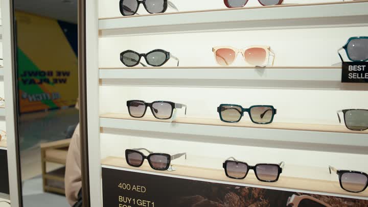 A collection of sunglasses in a Gulf store, purchasing sun protection glasses, a close-up shot from the side of an Arab Gulf Emirati woman wearing a colorful abaya choosing sunglasses from the displayed shelves, a Saudi woman shopping inside the mall