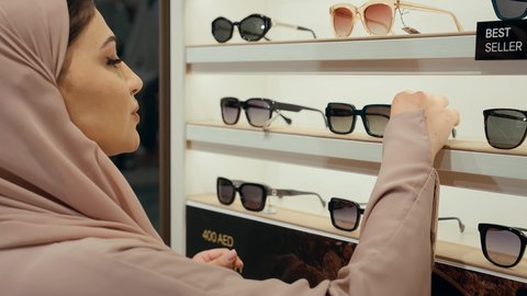 تشكيلة واسعة جديدة من نظارات النظر في محل خليجي، اقتناء نظارات حماية من الشمس، تصوير مقرب من الخلف لامراة عربيه خليجيه اماراتية ترتدي العباءه الملونه تقوم باختيار نظاره شمسيه من الارفف المعروضه، سيده سعوديه تقوم بالتسوق داخل المول
