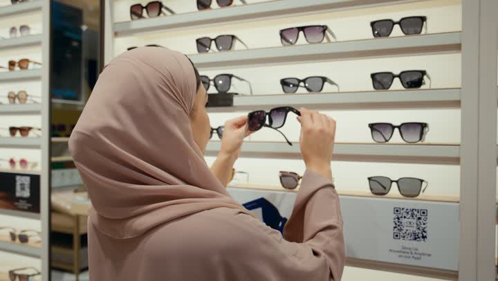 A new wide range of eyeglasses in a Gulf store, purchasing sunglasses, a close-up shot from the back of an Arab Gulf Emirati woman wearing a colorful abaya choosing sunglasses from the displayed shelves, a Saudi woman shopping inside the mall