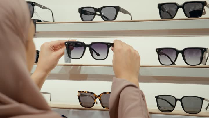 A new wide range of eyeglasses in a Gulf store, purchasing sunglasses, a close-up shot from the back of an Arab Gulf Emirati woman wearing a colorful abaya choosing sunglasses from the displayed shelves, a Saudi woman shopping inside the mall