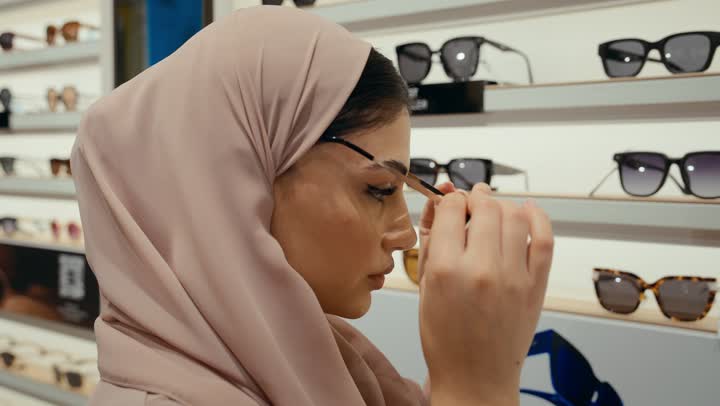 A Saudi woman is shopping inside the mall, buying sunglasses, a wide new selection of eyeglasses in a Gulf store, a close-up of an Arab Gulf Emirati woman wearing a colorful abaya choosing sunglasses from the displayed shelves, a Saudi woman is shopping inside the mall