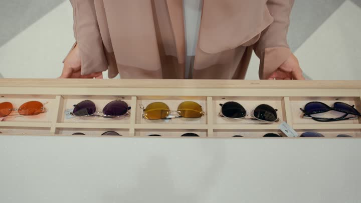 Buying sunglasses, a new wide range of eyeglasses in a Gulf store, how to choose sunglasses that suit your facial features, a close-up shot from the front of an Emirati Gulf Arab woman wearing a colorful abaya choosing sunglasses from inside the drawers, a Saudi woman shopping inside the mall