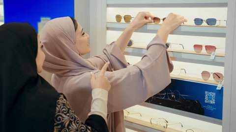كيفية إختيار النظارة الشمسية الملائمة لملامح الوجه ، اقتناء نظارات حماية من الشمس، تصوير مقرب لامرأتان عربيتان خليجيتان اماراتيان ترتديان الحجاب و العباءة تتشاركان لحظه مرحه اثناء فحص اطار نظارة شمسيه