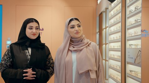 تبادل الراي و المشاركه في التسوق،تسوق و اختيار النظارات داخل متجر بصريات، امرأتان عربيتان خليجيتان اماراتيان ترتديان الحجاب و العباءة تتشاركان لحظه مرحه اثناء فحص اطار نظارة طبية