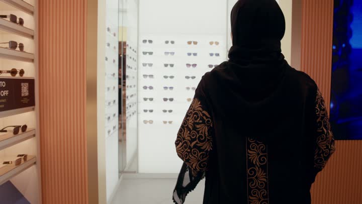 اقتناء نظارات حماية من الشمس، كيفية إختيار النظارة الشمسية الملائمة لملامح الوجه , تشكيلة نظارات شمسية أنيقة ، تصوير من الخلف لامرأة عربية خليجيه اماراتية ترتدي العباءه السوداء تقوم بدخول محل لاختيار الشكل المناسب