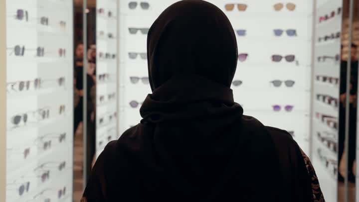 كيفية إختيار النظارة الشمسية الملائمة لملامح الوجه , تشكيلة نظارات شمسية أنيقة ، اقتناء نظارات حماية من الشمس، تصوير من الخلف لامرأة عربية خليجيه اماراتية ترتدي العباءه السوداء تقوم بدخول محل لاختيار الشكل المناسب