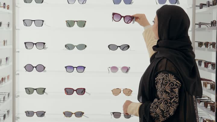 ارتداء و اختيار النظاره الشمسيه، امرأة عربية خليجيه اماراتية ترتدي العباءه السوداء تقوم بقياس النظارات لاختيار المناسب في محل نظارات , فتاة سعودية تلقي نظرة على مجموعة من الاطارات داخل المول