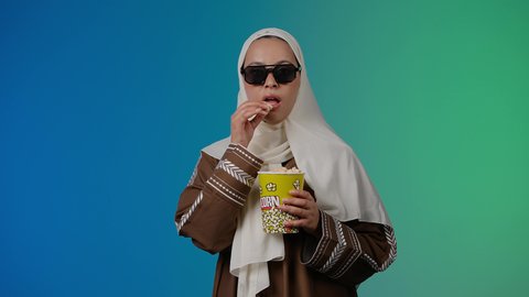 متابعة الفيلم المفضل ، تعابير السرور والسعاده، الاستمتاع باكل البوب كورن ، مشاهدة الأفلام داخل السينما، التسلية والترفيه بقضاء أوقات ممتعة،  تصوير مقرب لامرأة عربية خليجية اماراتيه محجبة ترتدي عباءه وتحمل علبه فوشار ، خلفية ملونه