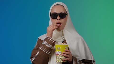 تعابير السرور والسعاده، الاستمتاع باكل البوب كورن ، متابعة الفيلم المفضل ، مشاهدة الأفلام داخل السينما، التسلية والترفيه بقضاء أوقات ممتعة،  تصوير مقرب لامرأة عربية خليجية اماراتيه محجبة ترتدي عباءه وتحمل علبه فوشار ، خلفية ملونه