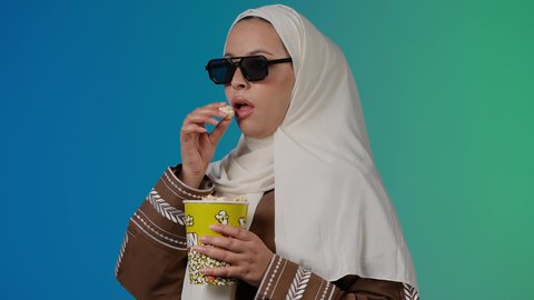 الاستمتاع باكل البوب كورن ، متابعة الفيلم المفضل ، مشاهدة الأفلام داخل السينما،التسلية والترفيه بقضاء أوقات ممتعة،  تصوير مقرب لامرأة عربية خليجية اماراتيه محجبة ترتدي عباءه وتحمل علبه فوشار ، خلفية ملونه