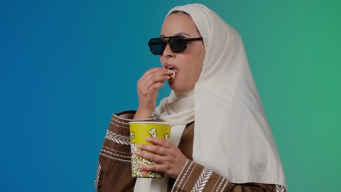 متابعة الفيلم المفضل ، مشاهدة الأفلام داخل السينما،التسلية والترفيه بقضاء أوقات ممتعة، الاستمتاع باكل البوب كورن ،  تصوير مقرب لامرأة عربية خليجية اماراتيه محجبة ترتدي عباءه وتحمل علبه فوشار ، خلفية ملونه