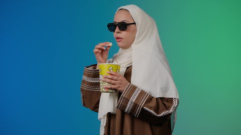 مشاهدة الأفلام داخل السينما،التسلية والترفيه بقضاء أوقات ممتعة، متابعة الفيلم المفضل ، الاستمتاع باكل البوب كورن ،  تصوير مقرب لامرأة عربية خليجية اماراتيه محجبة ترتدي عباءه وتحمل علبه فوشار ، خلفية ملونه