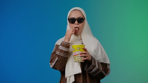 النظر بايماءات التفكير فى شئ ما ،تعابير الحيره والتأمل ، التسلية والترفيه بقضاء أوقات ممتعة، متابعة الفيلم المفضل ، الاستمتاع باكل البوب كورن ، مشاهدة الأفلام داخل السينما، تصوير مقرب لامرأة عربية خليجية اماراتيه محجبة ترتدي عباءه وتحمل علبه فوشار ، خلفية ملونه