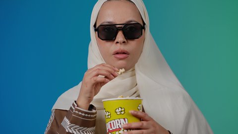 تعابير الحيره والتأمل ،النظر بايماءات التفكير فى شئ ما ، التسلية والترفيه بقضاء أوقات ممتعة، متابعة الفيلم المفضل ، الاستمتاع باكل البوب كورن ، مشاهدة الأفلام داخل السينما، تصوير مقرب لامرأة عربية خليجية اماراتيه محجبة ترتدي عباءه وتحمل علبه فوشار ، خلفية ملونه