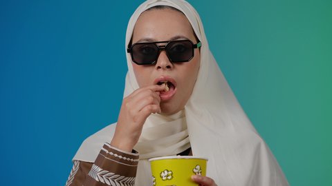 التسلية والترفيه بقضاء أوقات ممتعة، النظر بايماءات المفاجأه ،متابعة الفيلم المفضل ، تعابير الصدمه والذهول ، الاستمتاع باكل البوب كورن ، مشاهدة الأفلام داخل السينما، تصوير مقرب لامرأة عربية خليجية اماراتيه محجبة ترتدي عباءه وتحمل علبه فوشار ، خلفية ملونه
