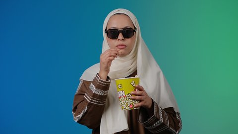 متابعة الفيلم المفضل ،رفع اليد بايماءات الدهشه ، تعابير الصدمه والذهول ، الاستمتاع باكل البوب كورن ، مشاهدة الأفلام داخل السينما، التسلية والترفيه بقضاء أوقات ممتعة، تصوير مقرب لامرأة عربية خليجية اماراتيه محجبة ترتدي عباءه وتحمل علبه فوشار ، خلفية ملونه