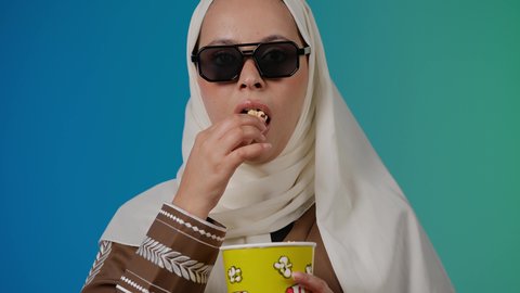 رفع اليد بايماءات الدهشه ، تعابير الصدمه والذهول ، الاستمتاع باكل البوب كورن ، مشاهدة الأفلام داخل السينما، التسلية والترفيه بقضاء أوقات ممتعة، متابعة الفيلم المفضل ، تصوير مقرب لامرأة عربية خليجية اماراتيه محجبة ترتدي عباءه وتحمل علبه فوشار ، خلفية ملونه