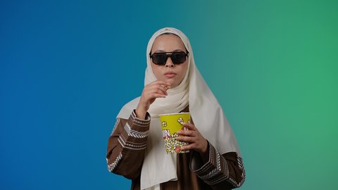 تعابير الموافقه والرضي، الاستمتاع باكل البوب كورن ، مشاهدة الأفلام داخل السينما، التسلية والترفيه بقضاء أوقات ممتعة، متابعة الفيلم المفضل ، تصوير مقرب لامرأة عربية خليجية اماراتيه محجبة ترتدي عباءه وتحمل علبه فوشار ، خلفية ملونه