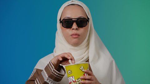 الاستمتاع باكل البوب كورن ، تعابير الموافقه والرضي، مشاهدة الأفلام داخل السينما، التسلية والترفيه بقضاء أوقات ممتعة، متابعة الفيلم المفضل ، تصوير مقرب لامرأة عربية خليجية اماراتيه محجبة ترتدي عباءه وتحمل علبه فوشار ، خلفية ملونه