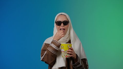 تحديد هدف معين ، الاشاره الى شئ ما، مشاهدة الأفلام داخل السينما، التسلية والترفيه بقضاء أوقات ممتعة، متابعة الفيلم المفضل ، الاستمتاع باكل البوب كورن وشرب المشروبات البارده ، تصوير مقرب لامرأة عربية خليجية اماراتيه محجبة ترتدي عباءه وتحمل علبه فوشار ، خلفية ملونه