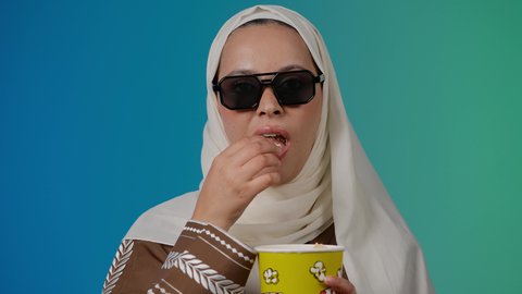 الاشاره الى شئ ما، مشاهدة الأفلام داخل السينما، التسلية والترفيه بقضاء أوقات ممتعة، متابعة الفيلم المفضل ، الاستمتاع باكل البوب كورن وشرب المشروبات البارده ، تصوير مقرب لامرأة عربية خليجية اماراتيه محجبة ترتدي عباءه وتحمل علبه فوشار ، خلفية ملونه