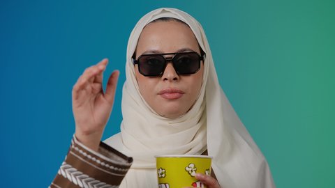 طلب التوقف عن فعل شيء ما،تعابير الحزم والنهي، مشاهدة الأفلام داخل السينما، التسلية والترفيه بقضاء أوقات ممتعة، متابعة الفيلم المفضل ، الاستمتاع باكل البوب كورن وشرب المشروبات البارده ، تصوير مقرب لامرأة عربية خليجية اماراتيه محجبة ترتدي عباءه وتحمل علبه فوشار ، خلفية ملونه