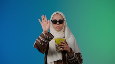 تعابير الحزم والنهي، طلب التوقف عن فعل شيء ما، مشاهدة الأفلام داخل السينما، التسلية والترفيه بقضاء أوقات ممتعة، متابعة الفيلم المفضل ، الاستمتاع باكل البوب كورن وشرب المشروبات البارده ، تصوير مقرب لامرأة عربية خليجية اماراتيه محجبة ترتدي عباءه وتحمل علبه فوشار ، خلفية ملونه