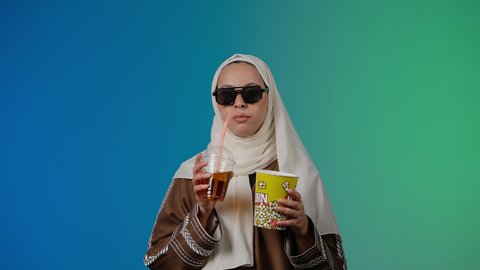 مشاهدة الأفلام داخل السينما، التسلية والترفيه بقضاء أوقات ممتعة، متابعة الفيلم المفضل بتعابير المرح والاستمتاع ، الاستمتاع  باكل البوب كورن وشرب المشروبات البارده ، تصوير مقرب لامرأة عربية خليجية اماراتيه محجبة ترتدي عباءه وتحمل علبه فوشار ، خلفية ملونه