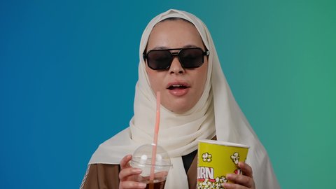 التسلية والترفيه بقضاء أوقات ممتعة، متابعة الفيلم المفضل بتعابير المرح والاستمتاع ، الاستمتاع  باكل البوب كورن وشرب المشروبات البارده ، مشاهدة الأفلام داخل السينما، تصوير مقرب لامرأة عربية خليجية اماراتيه محجبة ترتدي عباءه وتحمل علبه فوشار ، خلفية ملونه
