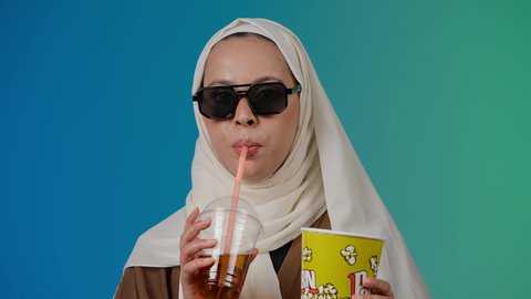 متابعة الفيلم المفضل بتعابير المرح والاستمتاع ، الاستمتاع  باكل البوب كورن وشرب المشروبات البارده ،  التسلية والترفيه بقضاء أوقات ممتعة، مشاهدة الأفلام داخل السينما، تصوير مقرب لامرأة عربية خليجية اماراتيه محجبة ترتدي عباءه وتحمل علبه فوشار ، خلفية ملونه