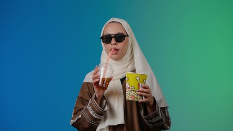 الاستمتاع  باكل البوب كورن وشرب المشروبات البارده ، ايماءات الجودة والامتياز والرضي ، التسلية والترفيه بقضاء أوقات ممتعة، مشاهدة الأفلام داخل السينما، متابعة الفيلم المفضل، تصوير مقرب لامرأة عربية خليجية اماراتيه محجبة ترتدي عباءه وتحمل علبه فوشار ، خلفية ملونه