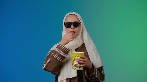 ايماءات الجودة والامتياز والرضي ، التسلية والترفيه بقضاء أوقات ممتعة، الاستمتاع بمشاهدة الأفلام داخل السينما، اكل البوب كورن ، متابعة الفيلم المفضل، تصوير مقرب لامرأة عربية خليجية اماراتيه محجبة ترتدي عباءه وتحمل علبه فوشار ، خلفية ملونه