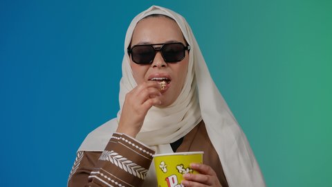 تعابير الموافقه والرضي ، الاستمتاع بمشاهدة الأفلام داخل السينما، اكل البوب كورن ، التسلية والترفيه بقضاء أوقات ممتعة، متابعة الفيلم المفضل، تصوير مقرب لامرأة عربية خليجية اماراتيه محجبة ترتدي عباءه وتحمل علبه فوشار ، خلفية ملونه