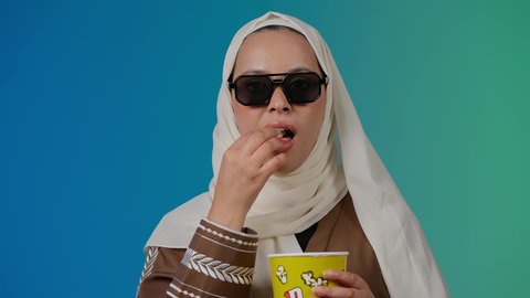 الاستمتاع بمشاهدة الأفلام داخل السينما، تعابير الحماس والسعاده ، اكل البوب كورن ، التسلية والترفيه بقضاء أوقات ممتعة، متابعة الفيلم المفضل، تصوير مقرب لامرأة عربية خليجية اماراتيه محجبة ترتدي عباءه وتحمل علبه فوشار ، ترفع قبضه اليد بايماءات السرور ، خلفية ملونه
