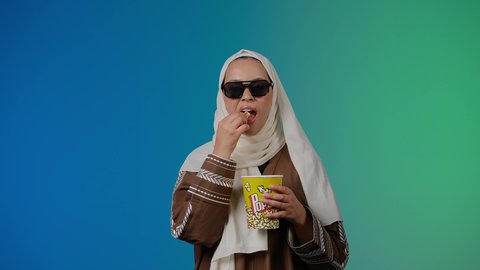 تعابير الحماس والسعاده ، اكل البوب كورن ، الاستمتاع بمشاهدة الأفلام داخل السينما، التسلية والترفيه بقضاء أوقات ممتعة، متابعة الفيلم المفضل، تصوير مقرب لامرأة عربية خليجية اماراتيه محجبة ترتدي عباءه وتحمل علبه فوشار ، ترفع قبضه اليد بايماءات السرور ، خلفية ملونه