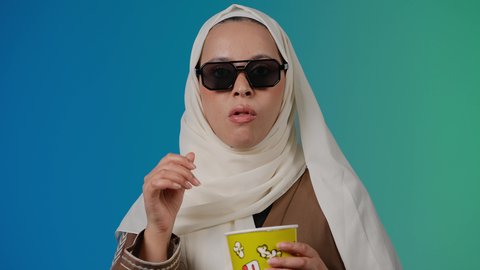 اكل البوب كورن ، الاستمتاع بمشاهدة الأفلام داخل السينما، التسلية والترفيه بقضاء أوقات ممتعة، متابعة الفيلم المفضل، تصوير مقرب لامرأة عربية خليجية اماراتيه محجبة ترتدي عباءه وتحمل علبه فوشار ، خلفية ملونه