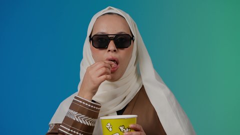 الاستمتاع بمشاهدة الأفلام داخل السينما، التسلية والترفيه بقضاء أوقات ممتعة، متابعة الفيلم المفضل، النظر من خلال نظاره سوداء ، اكل البوب كورن ، تصوير مقرب لامرأة عربية خليجية اماراتيه محجبة ترتدي عباءه وتحمل كيس فوشار ، خلفية ملونه