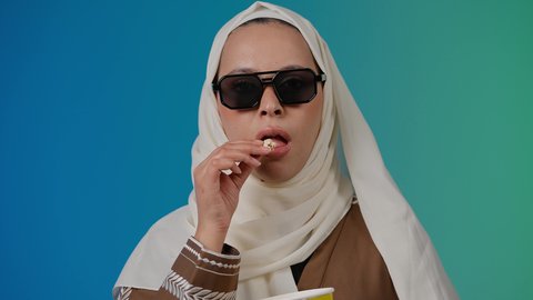 النظر من خلال نظاره سوداء ، الاستمتاع بمشاهدة الأفلام داخل السينما، اكل البوب كورن ، متابعة الفيلم المفضل، التسلية والترفيه بقضاء أوقات ممتعة، تصوير مقرب لامرأة عربية خليجية اماراتيه محجبة ترتدي عباءه وتحمل كيس فوشار ، خلفية ملونه