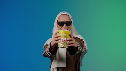 الاستمتاع بمشاهدة الأفلام داخل السينما، اكل البوب كورن ، متابعة الفيلم المفضل، التسلية والترفيه بقضاء أوقات ممتعة، تصوير مقرب لامرأة عربية خليجية اماراتيه محجبة ترتدي عباءه وتحمل كيس فوشار ، خلفية ملونه