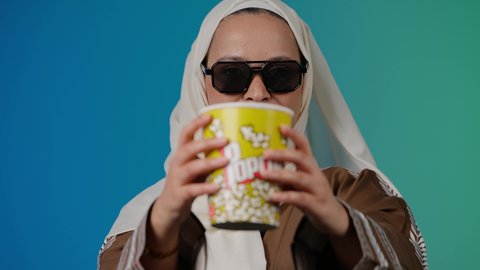 الاستمتاع باكل البوب كورن ، متابعة الفيلم المفضل، التسلية والترفيه بقضاء أوقات ممتعة، الاستمتاع بمشاهدة الأفلام داخل السينما، تصوير مقرب لامرأة عربية خليجية اماراتيه محجبة ترتدي عباءه وتحمل كيس فوشار ، خلفية ملونه