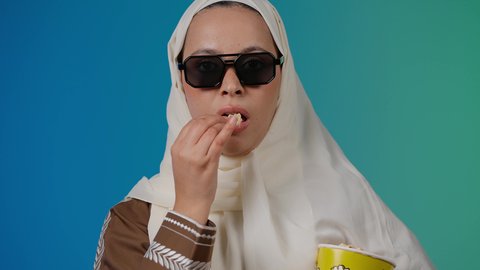 تعابير الخوف والانزعاج ، اكل البوب كورن ، متابعة الفيلم المفضل، التسلية والترفيه بقضاء أوقات ممتعة، الاستمتاع بمشاهدة الأفلام داخل السينما، تصوير مقرب لامرأة عربية خليجية اماراتيه محجبة ترتدي عباءه وتحمل كيس فوشار ، خلفية ملونه