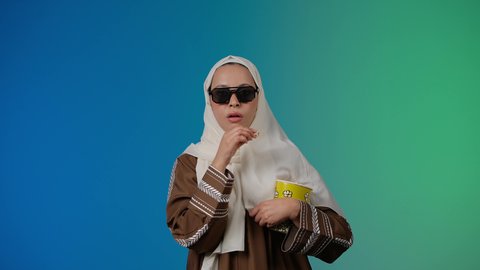 اكل البوب كورن ، متابعة الفيلم المفضل، التسلية والترفيه بقضاء أوقات ممتعة، تعابير السرور والسعاده ، الاستمتاع بمشاهدة الأفلام داخل السينما، تصوير مقرب لامرأة عربية خليجية اماراتيه محجبة ترتدي عباءه وتحمل كيس فوشار ، خلفية ملونه