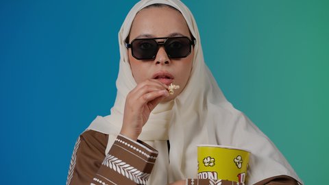 التسلية والترفيه بقضاء أوقات ممتعة، تعابير الاندماج والاستمتاع ،متابعة الفيلم المفضل، الاستمتاع بمشاهدة الأفلام داخل السينما، تصوير مقرب لامرأة عربية خليجية اماراتيه محجبة ترتدي عباءه وتحمل كيس فوشار ، خلفية ملونه