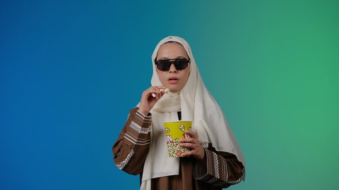 تعابير الاندماج والاستمتاع ،متابعة الفيلم المفضل، الاستمتاع بمشاهدة الأفلام داخل السينما،  التسلية والترفيه بقضاء أوقات ممتعة، تصوير مقرب لامرأة عربية خليجية اماراتيه محجبة ترتدي عباءه وتحمل كيس فوشار ، خلفية ملونه