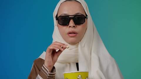 متابعة الفيلم المفضل، الاستمتاع بمشاهدة الأفلام داخل السينما، تعابير المرح والاستمتاع ، التسلية والترفيه بقضاء أوقات ممتعة، تصوير مقرب لامرأة عربية خليجية اماراتيه محجبة ترتدي عباءه وتحمل كيس فوشار  ، خلفية ملونه