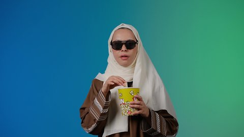 الاستمتاع بمشاهدة الأفلام داخل السينما، تعابير المرح والاستمتاع ، التسلية والترفيه بقضاء أوقات ممتعة، متابعة الفيلم المفضل، تصوير مقرب لامرأة عربية خليجية اماراتيه محجبة ترتدي عباءه وتحمل كيس فوشار والجوال ، خلفية ملونه