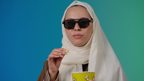 تعابير المرح والاستمتاع ، التسلية والترفيه بقضاء أوقات ممتعة، متابعة الفيلم المفضل، الاستمتاع بمشاهدة الأفلام داخل السينما، تصوير مقرب لامرأة عربية خليجية اماراتيه محجبة ترتدي عباءه وتحمل كيس فوشار والجوال ، خلفية ملونه