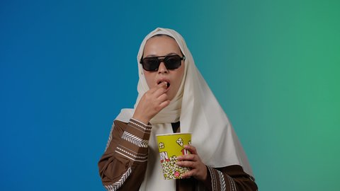 تعابير الخوف والرعب والصدمه ، الاستمتاع بمشاهدة الأفلام داخل السينما، اكل البوب كورن ، التسلية والترفيه بقضاء أوقات ممتعة، متابعة الفيلم المفضل، تصوير مقرب لامرأة عربية خليجية اماراتيه محجبة ترتدي عباءه وتحمل علبه فوشار ، خلفية ملونه