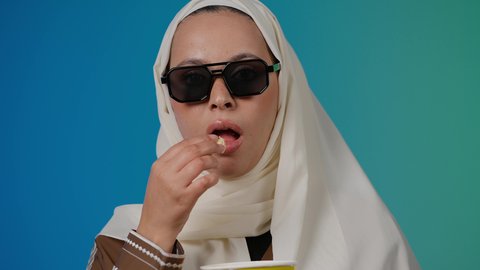 تعابير الذهول والمفاجأه ، الاستمتاع بمشاهدة الأفلام داخل السينما، اكل البوب كورن ، التسلية والترفيه بقضاء أوقات ممتعة، متابعة الفيلم المفضل، تصوير مقرب لامرأة عربية خليجية اماراتيه محجبة ترتدي عباءه وتحمل علبه فوشار ، خلفية ملونه